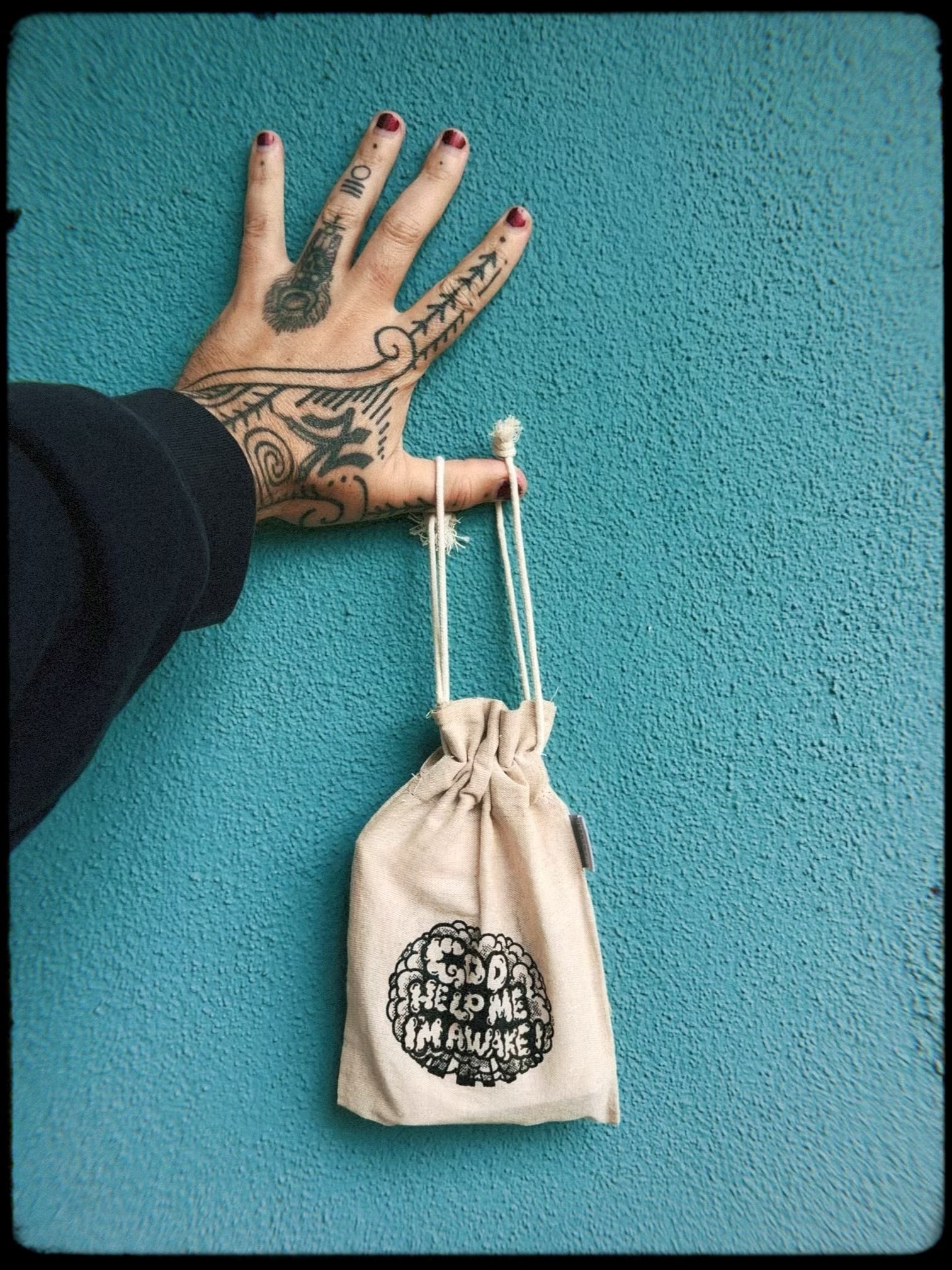 Tattooed hand dangling a burlap bag that says God Help Me, I'm Awake! 