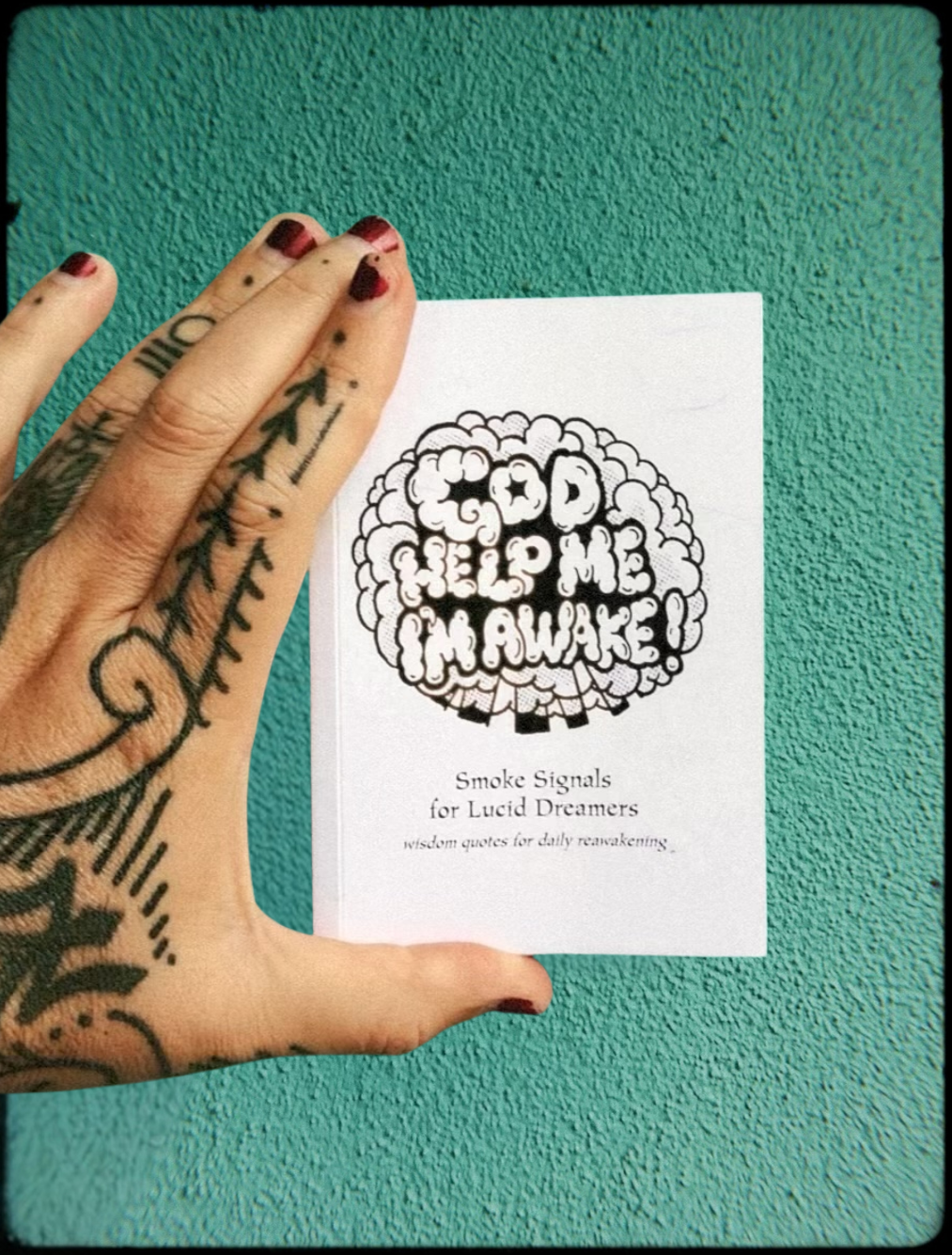 God Help Me, I'm Awake! (Volume 1) Wisdom Quotes for Daily Reawakening book being held by a tattooed hand on a turquoise background