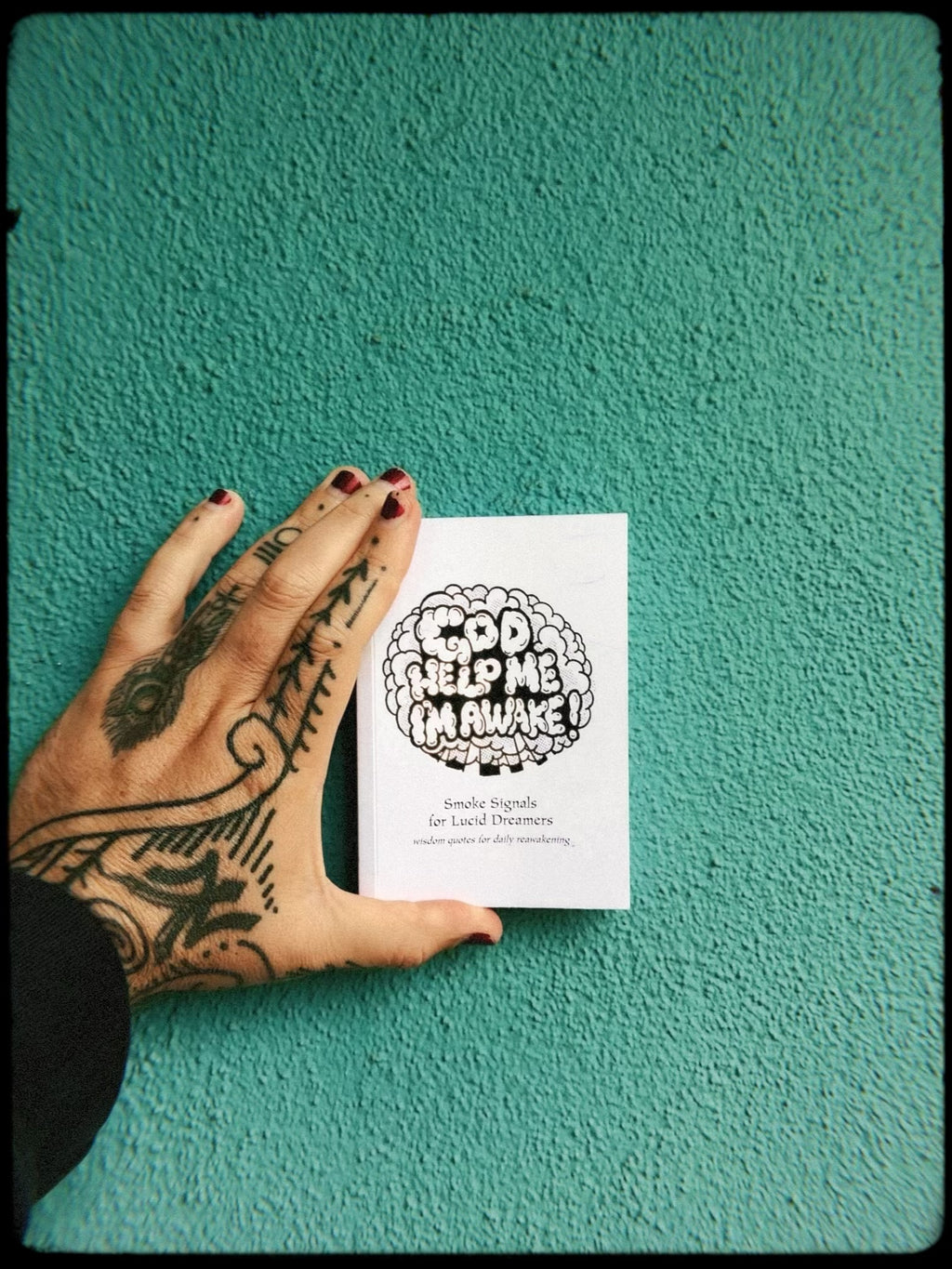 God Help Me, I'm Awake! (Volume 1) Wisdom Quotes for Daily Reawakening book being held by a tattooed hand on a turquoise background